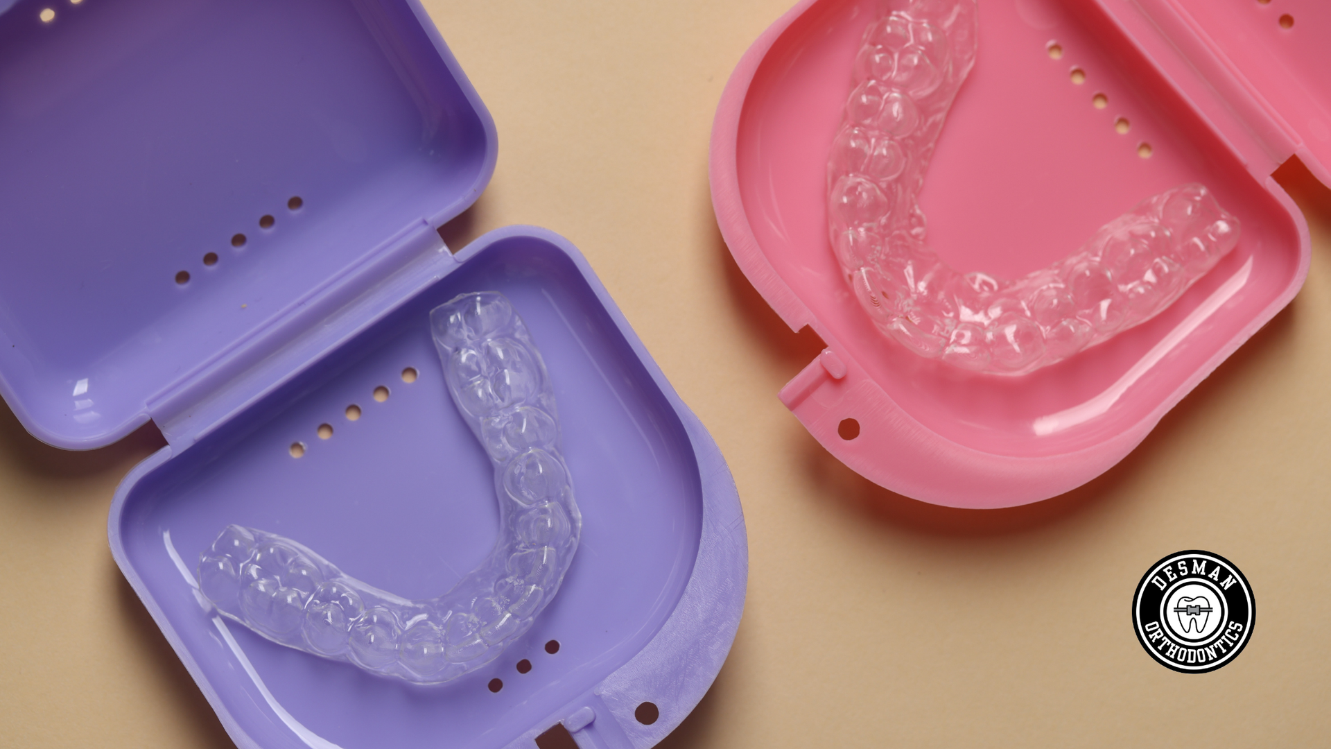 Open cases with clear dental aligners in purple and pink trays on a tan surface.