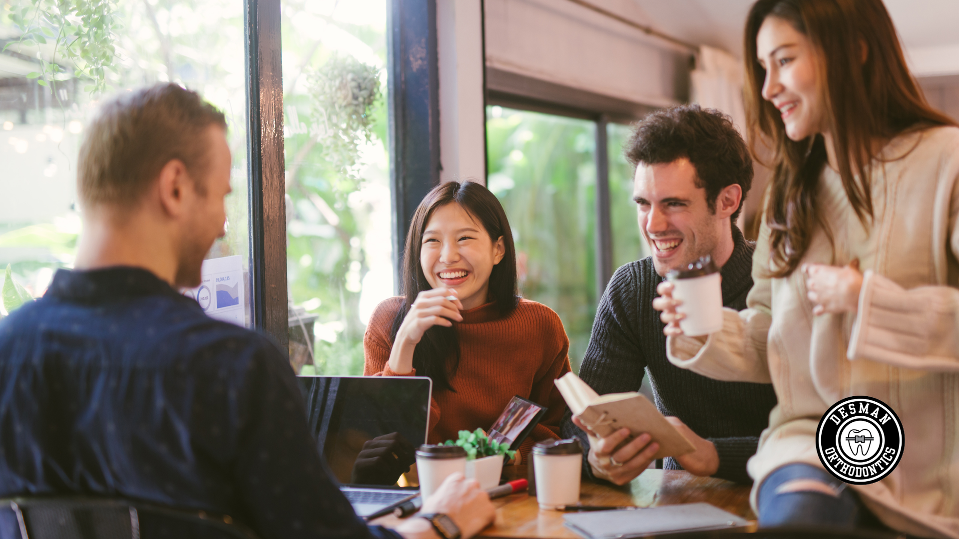 Friends chatting and smiling around a table in a bright cafe, with laptops and drinks.