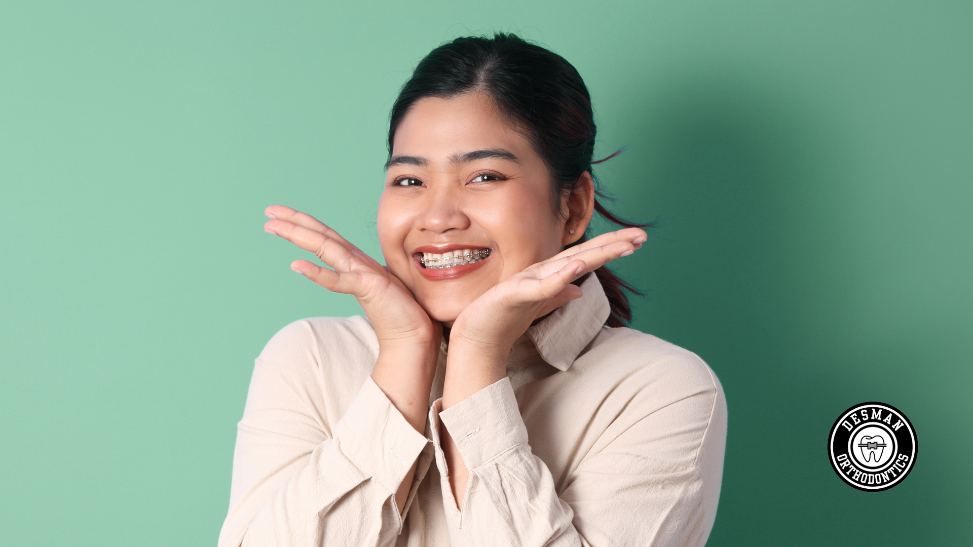 Smiling person posing with hands under chin against a mint green background.