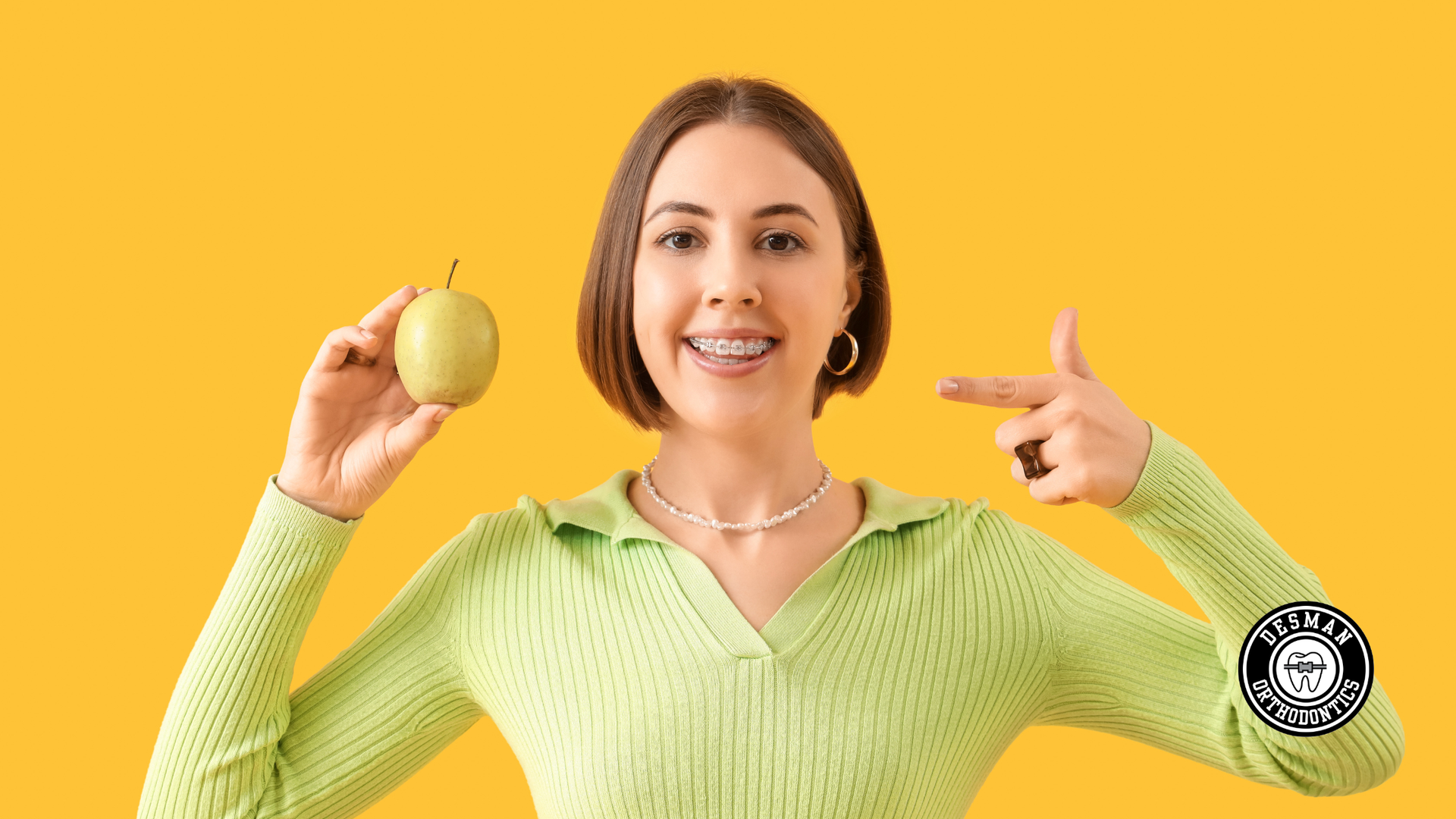 Smiling person in a light green sweater holding a green fruit and pointing at it against a yellow background