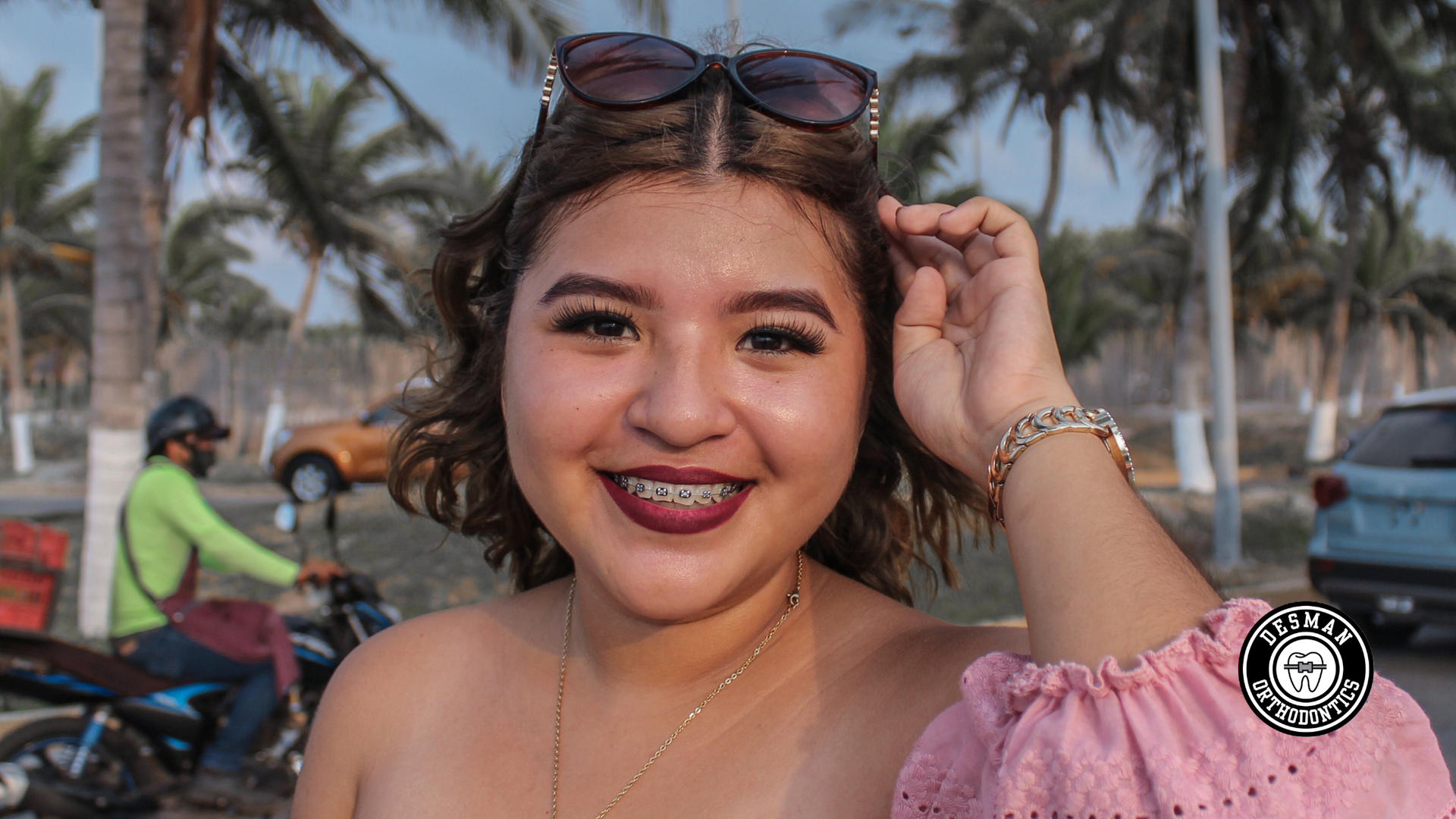 Smiling woman in pink off-shoulder top adjusts sunglasses at a palm-lined beach street