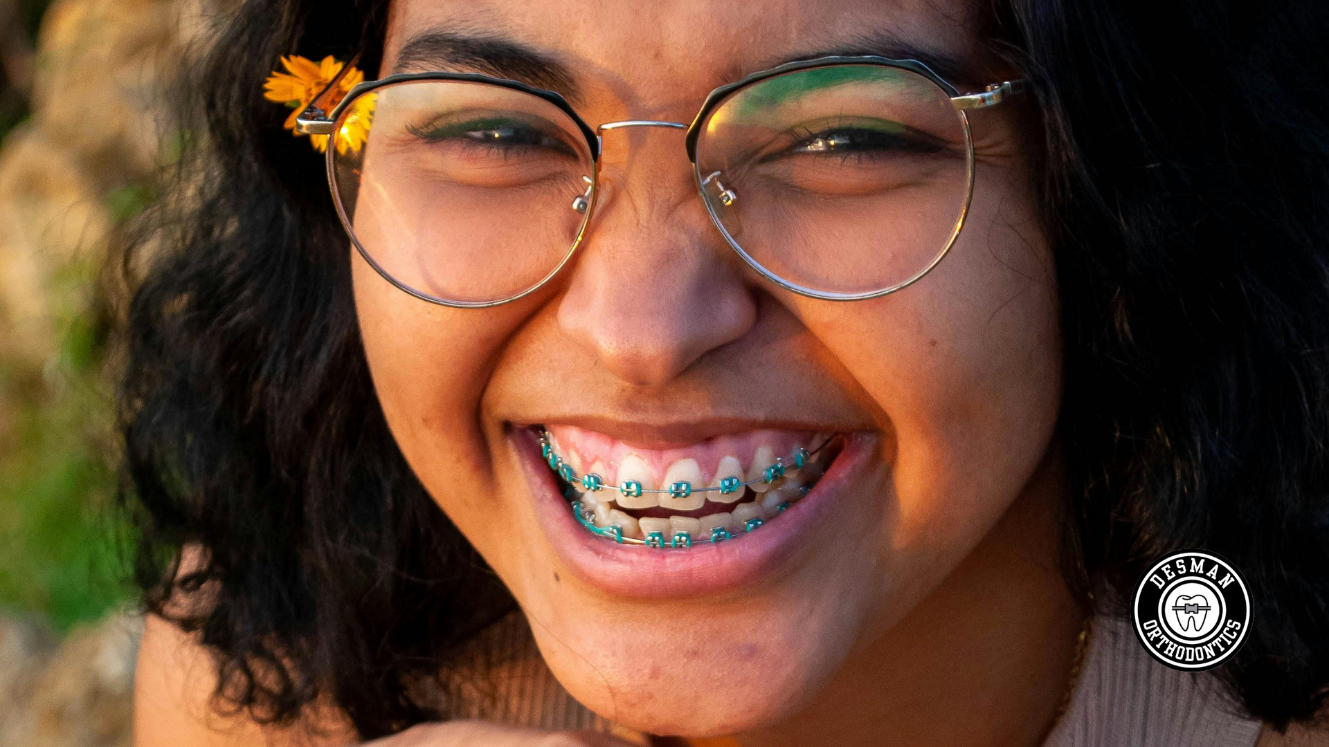 Smiling person with glasses and braces, wearing a flower in their hair