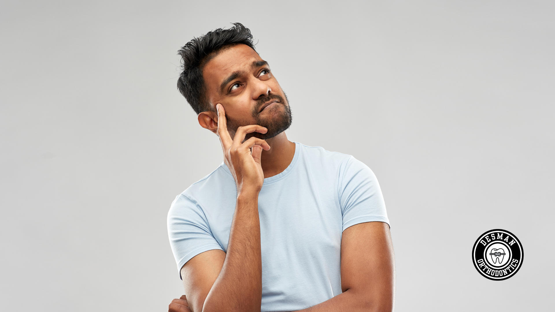 Man in a light blue shirt thoughtfully touching his chin against a plain gray background