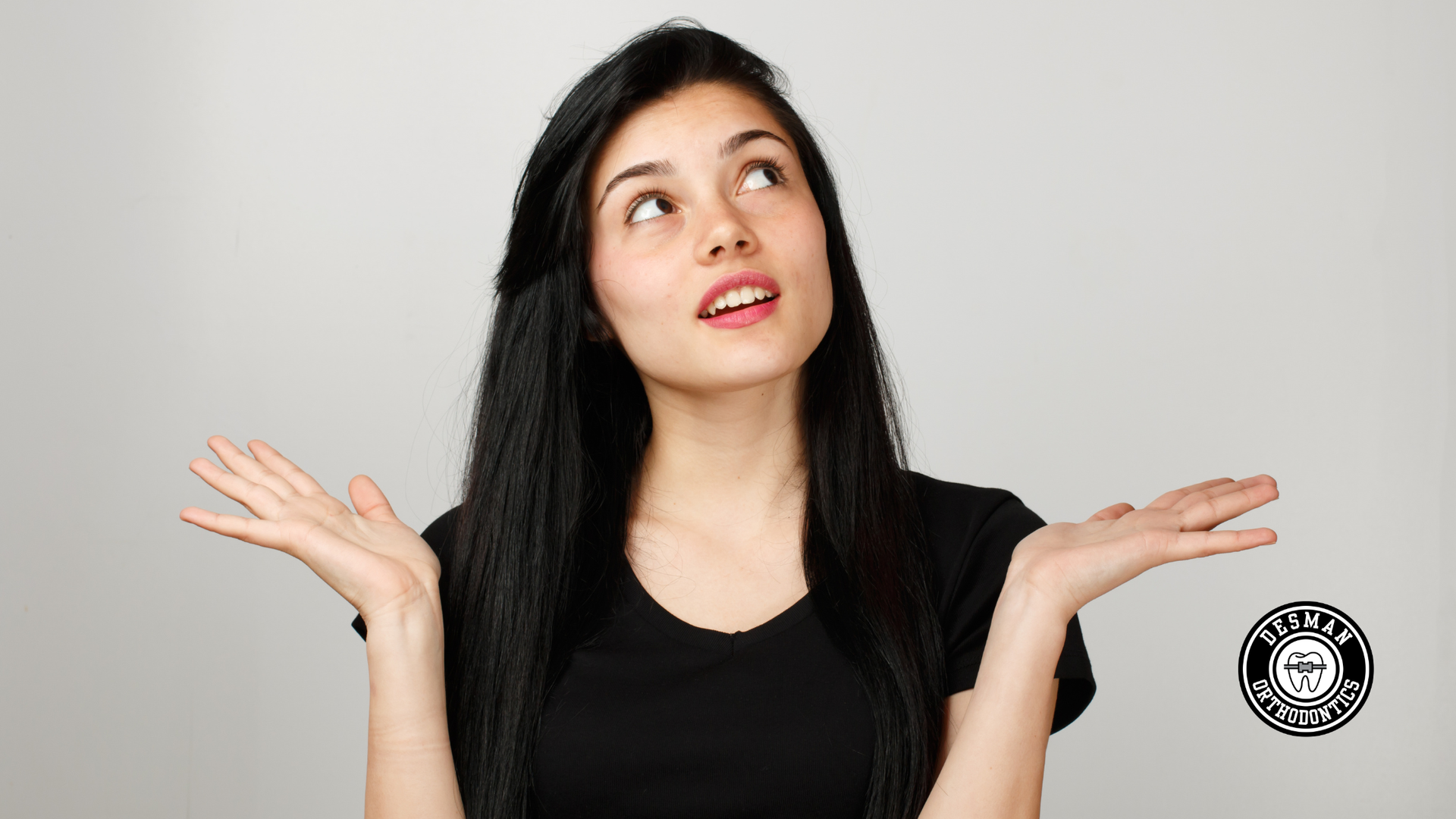 Woman with long black hair shrugging with palms up and looking upward against a light gray background