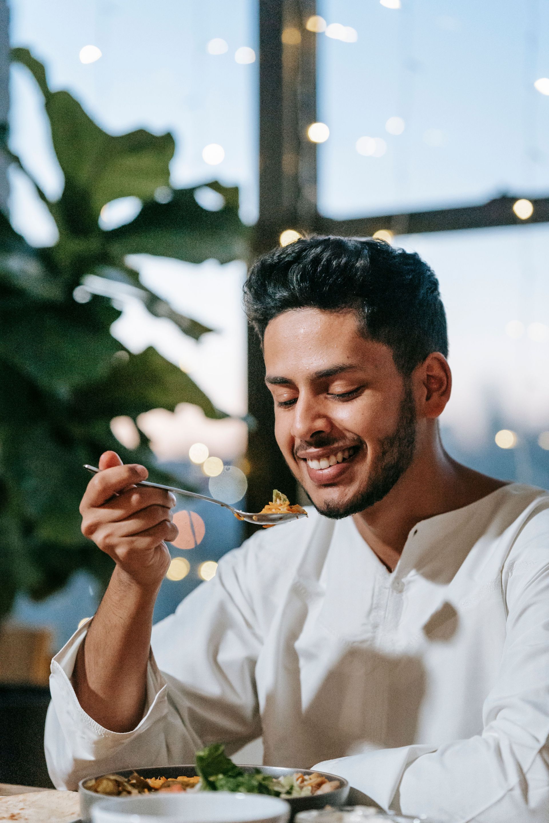 Smiling person in a white shirt eating at a table by a window with plants in the background