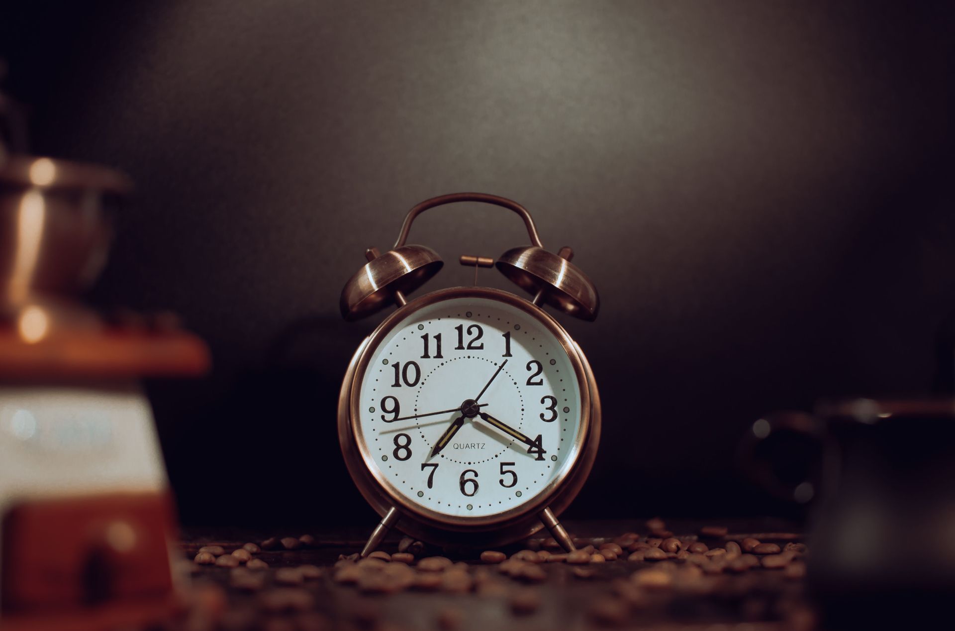 A vintage alarm clock on a tabletop, softly lit with blurred background objects.
