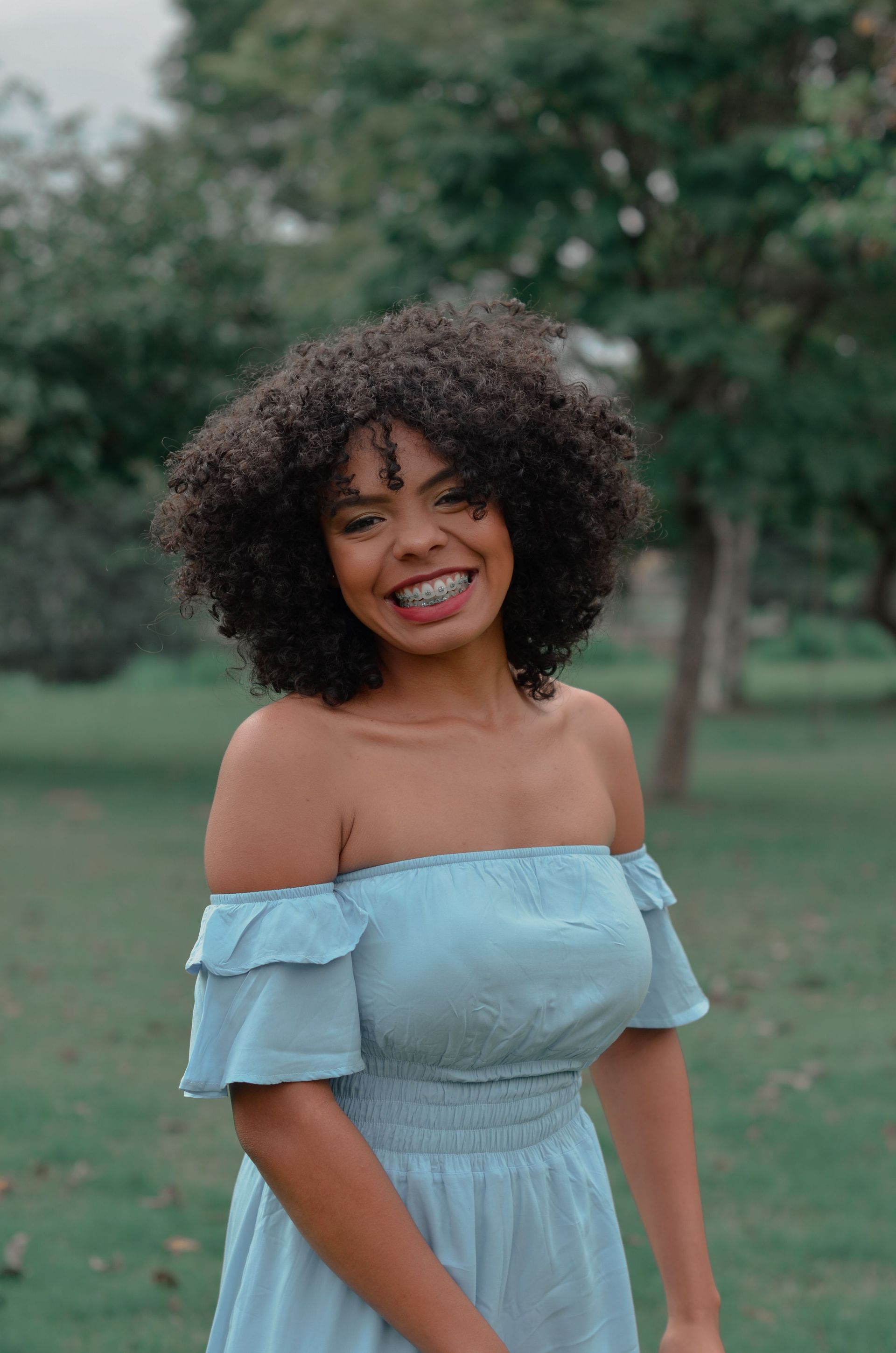 Smiling woman in a light blue off-shoulder dress standing outdoors in a grassy park.