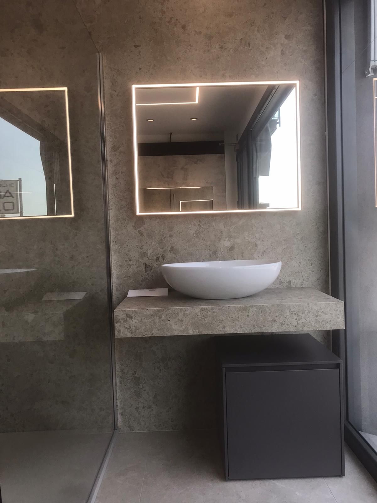 Mobile bagno moderno con lavabo rotondo bianco, specchio retroilluminato e mobiletto scuro sotto un piano in pietra.