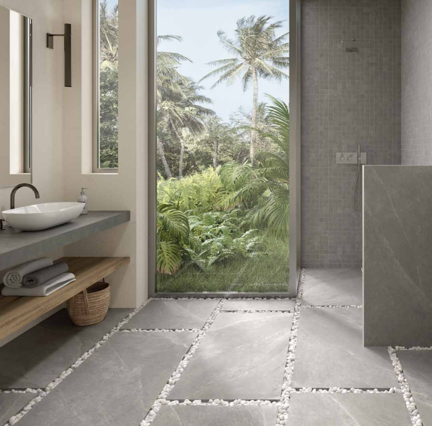 Bagno moderno con pareti in pietra grigia, lavabo sotto la finestra e porta a vetri che si apre su una vegetazione tropicale.