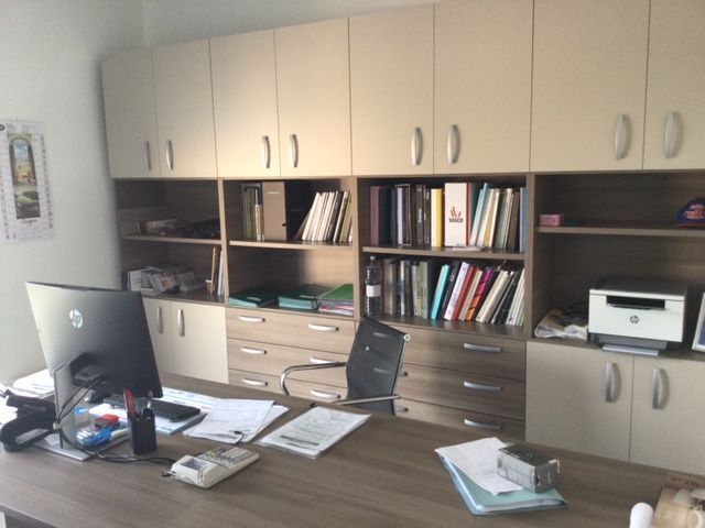 Ufficio con armadietti beige, scaffali pieni di libri e una scrivania ingombra con computer e documenti.