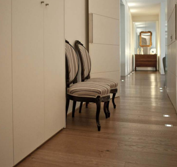 Luminoso corridoio con pareti beige, pavimento in legno, sedie a righe e uno specchio in fondo.
