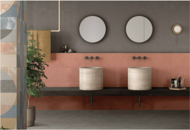 Bagno moderno con due specchi rotondi, doppio lavabo e un lungo piano d'appoggio in legno su pareti rosa e grigie.