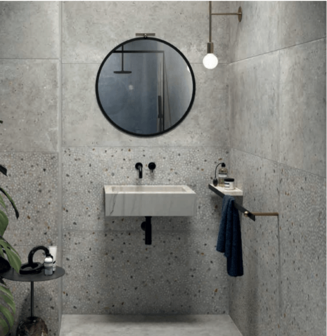 Bagno moderno di colore grigio con specchio rotondo, lavabo a parete e portasciugamani, illuminato da una lampadina sospesa.