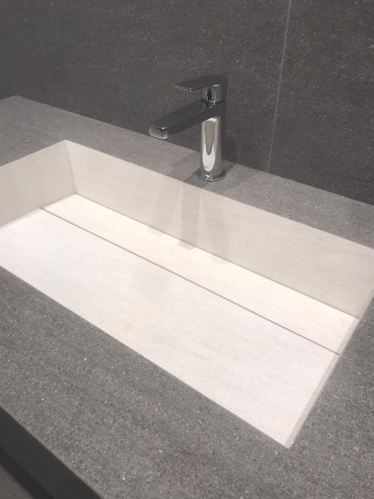 Lavabo moderno con vasca bianca e piano d'appoggio scuro su pareti rivestite di piastrelle grigie.