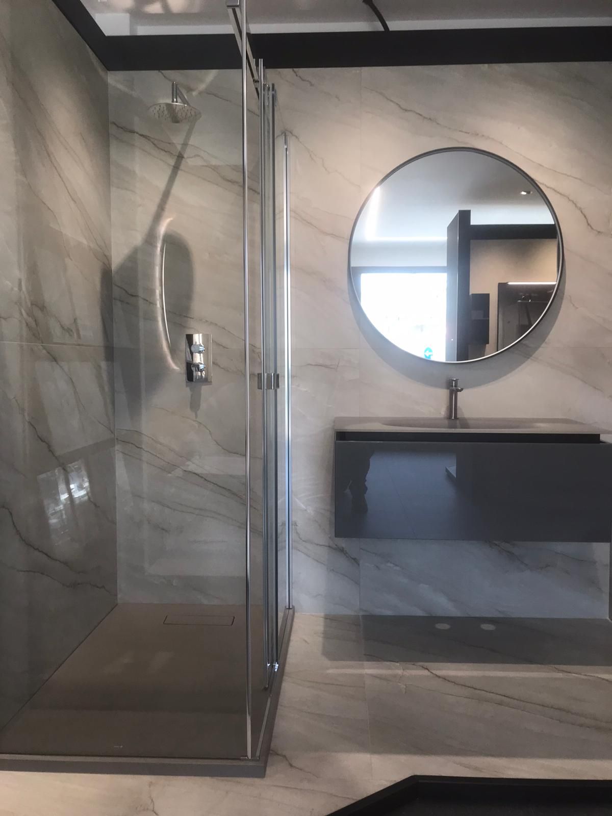 Bagno moderno con doccia in vetro, specchio rotondo, mobile lavabo sospeso nero e pavimento piastrellato.