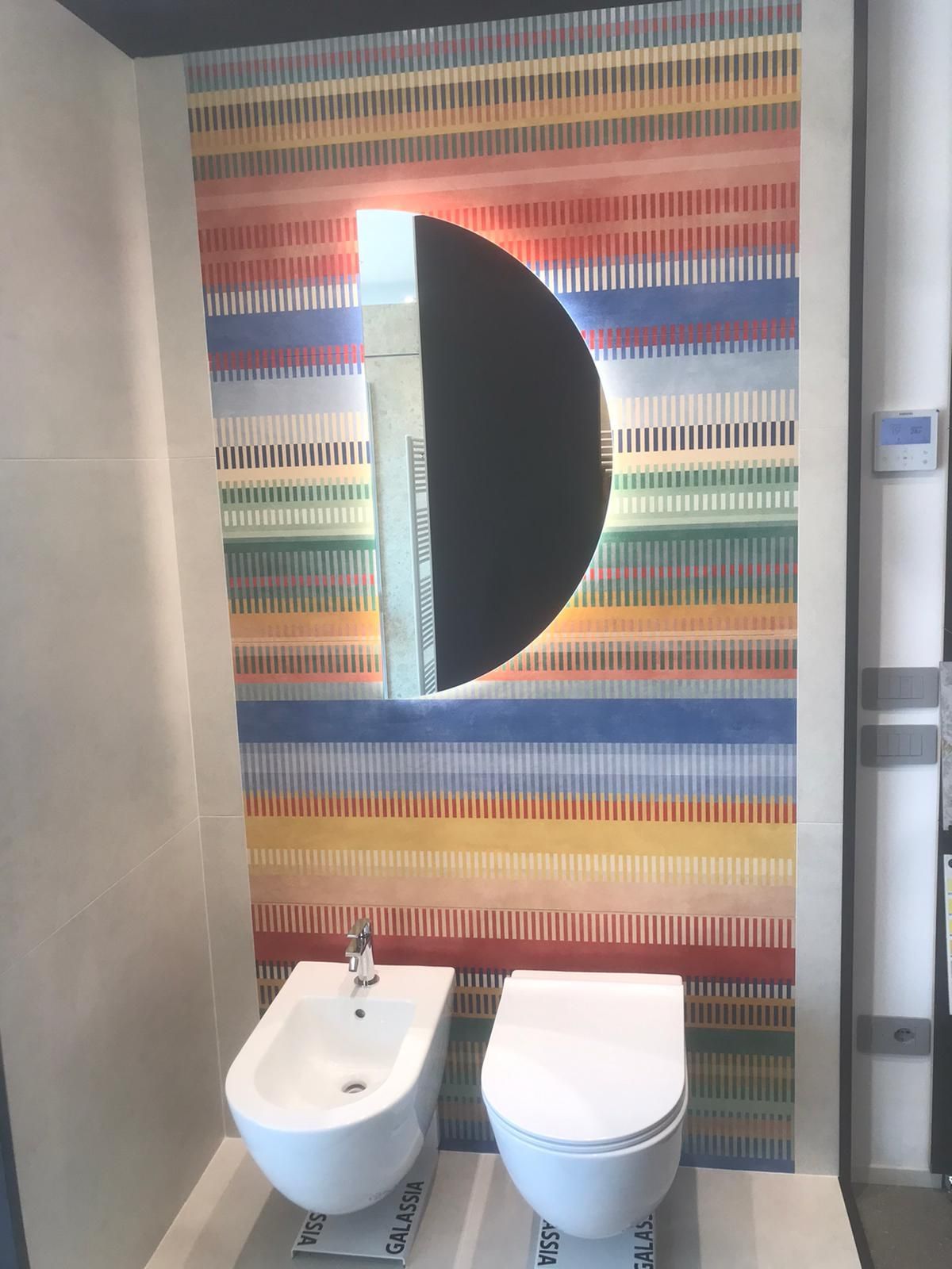 Bagno moderno con parete rivestita di piastrelle a righe colorate, specchio rotondo, lavabo e WC.