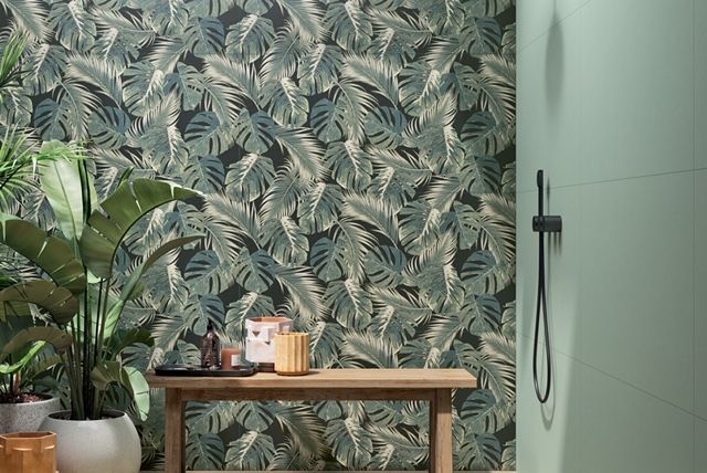 Carta da parati con foglie tropicali, pianta in vaso e tavolo in legno accanto a una cabina doccia con rubinetteria nera.