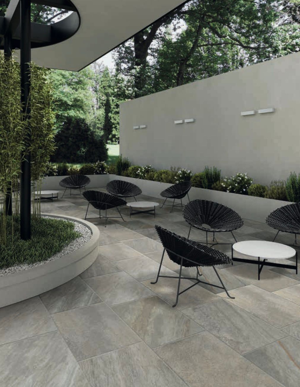 Moderno patio esterno con sedie a sdraio nere, tavolini rotondi e piante in vaso accanto a un muro di cemento.