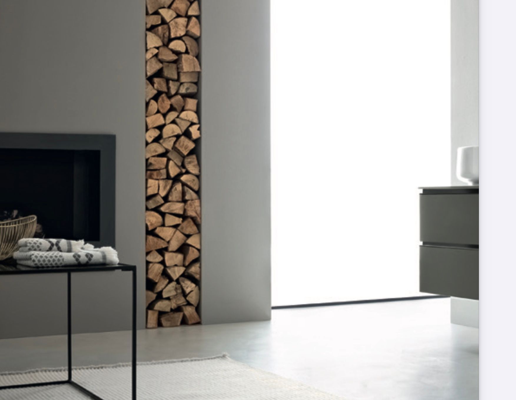 Soggiorno moderno e minimalista con legna da ardere accatastata, camino scuro e luce brillante proveniente dalla finestra.