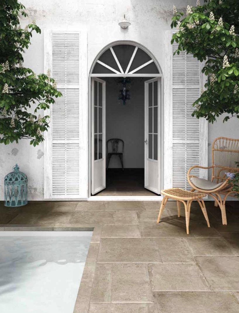 Patio con porte finestre bianche aperte, sedia in vimini, lanterna e piscina accanto a una casa bianca.