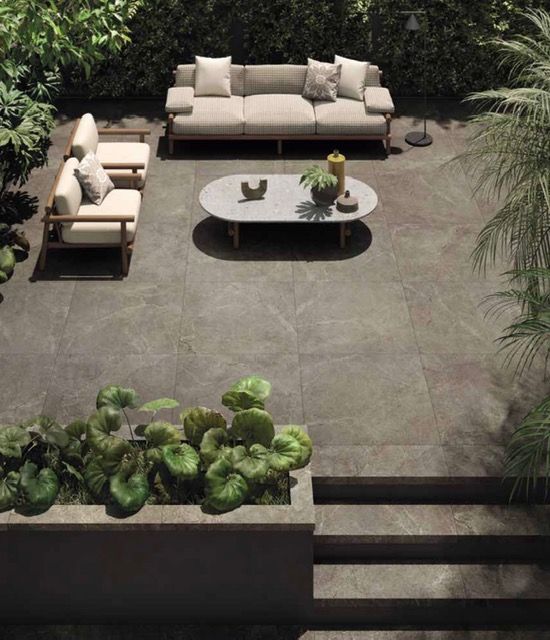 Patio moderno con sedie a sdraio beige, tavolino ovale e vegetazione lussureggiante su pavimentazione in pietra grigia.