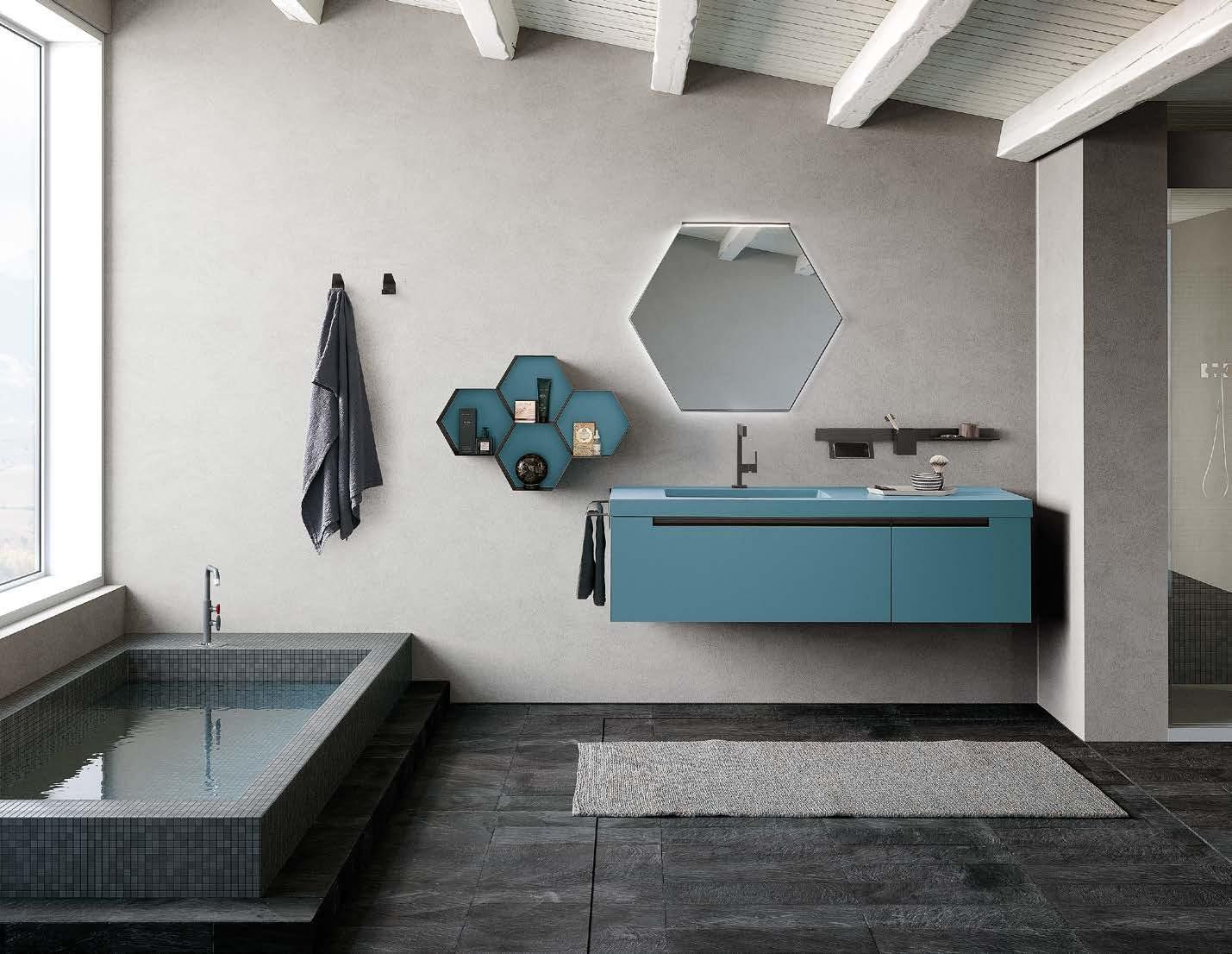 Bagno moderno con piastrelle grigie, mobile lavabo blu, specchio esagonale e vasca da bagno piastrellata accanto a una finestra.