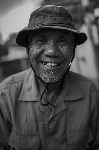 Smiling person in a dark bucket hat and button-up shirt, standing outdoors in black and white.