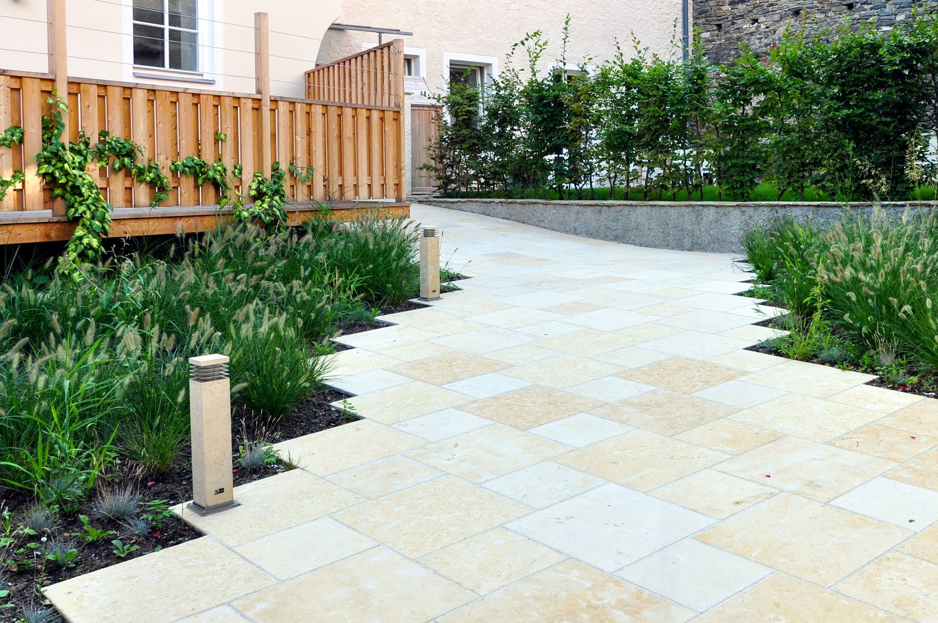 A paved stone walkway winds through a garden with green plants and low, modern light fixtures next to a wooden fence.