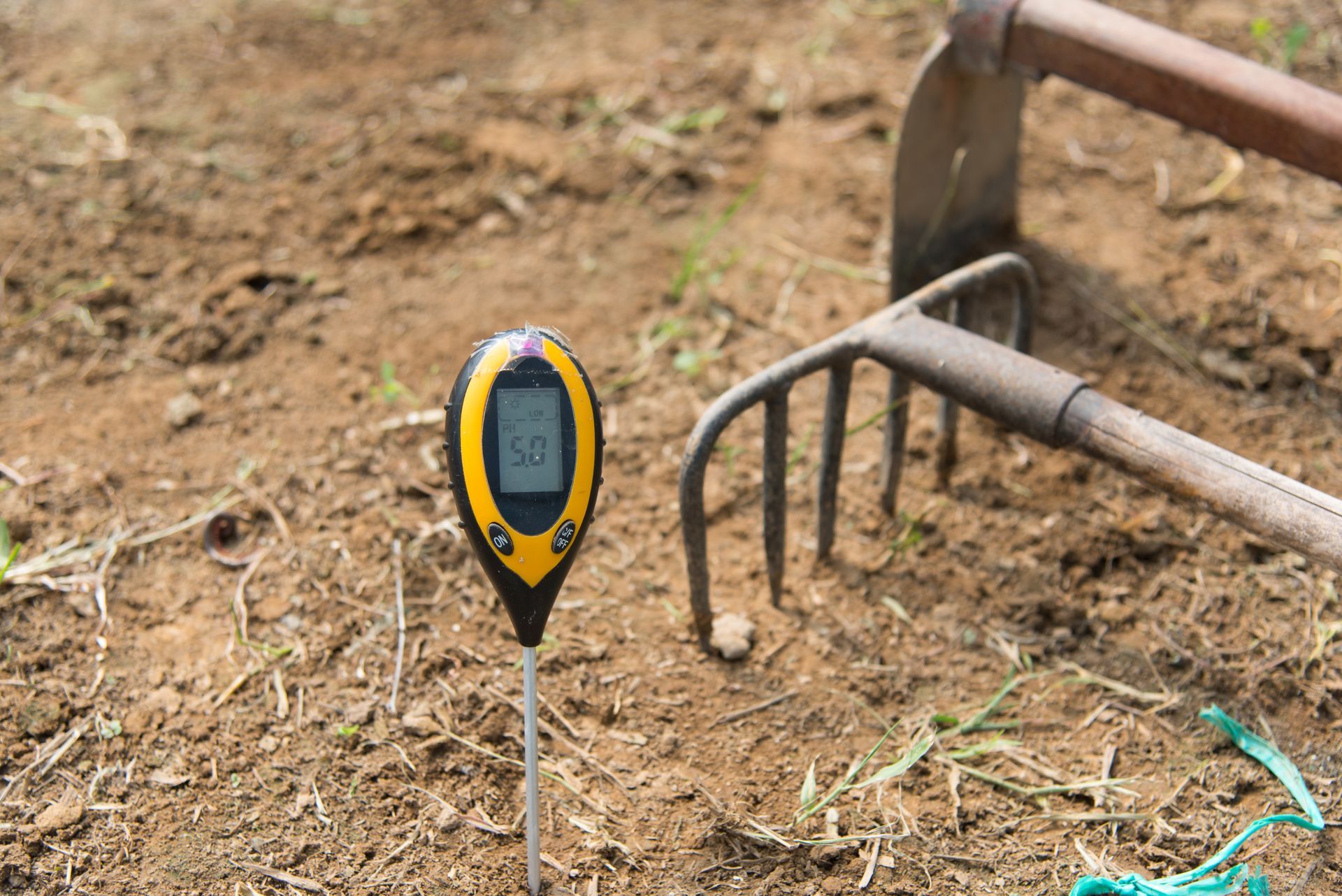 Digital soil moisture meter beside a metal garden rake on dry ground