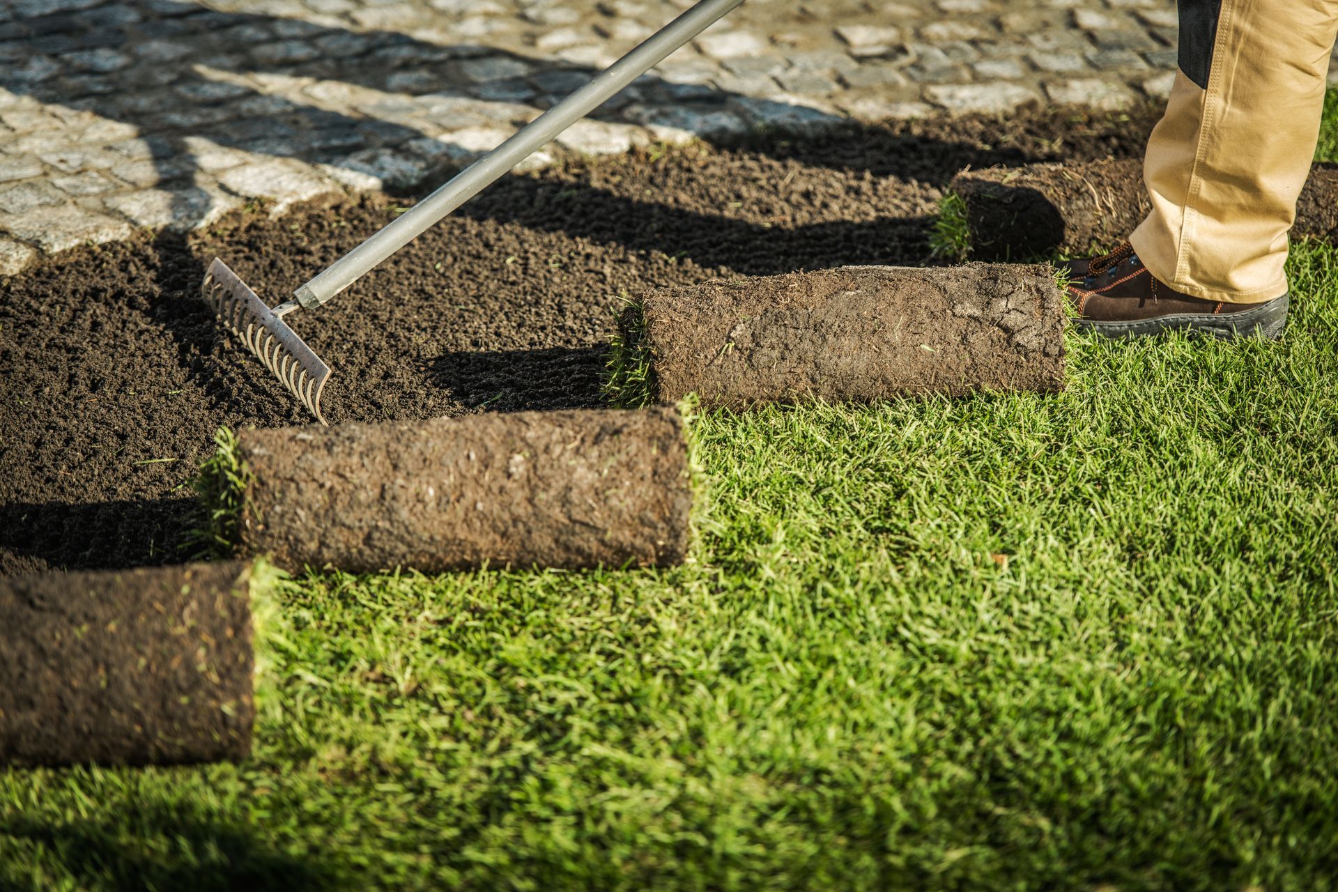 Sod being laid over soil with a rake on a cobblestone path beside a worker’s boot
