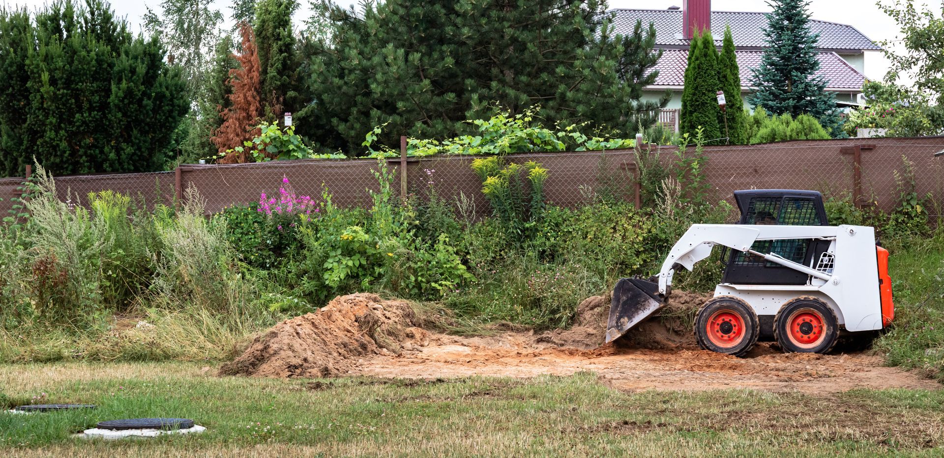 Small skid-steer loader pushing a dirt pile in a grassy lot with trees and a fence behind it
