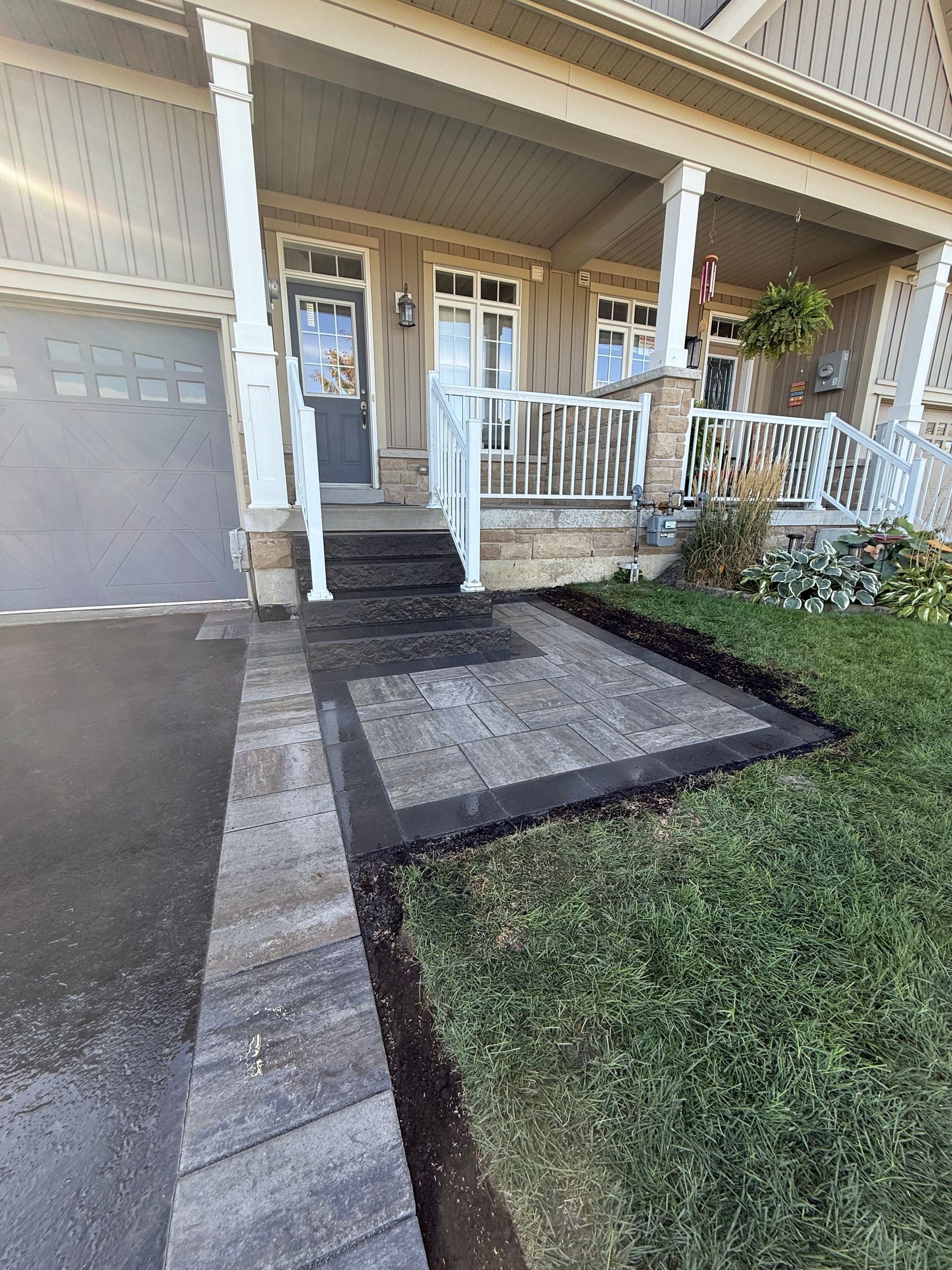 A home's entrance with a paved walkway and patio. Green grass borders the pavers. A covered porch is visible.