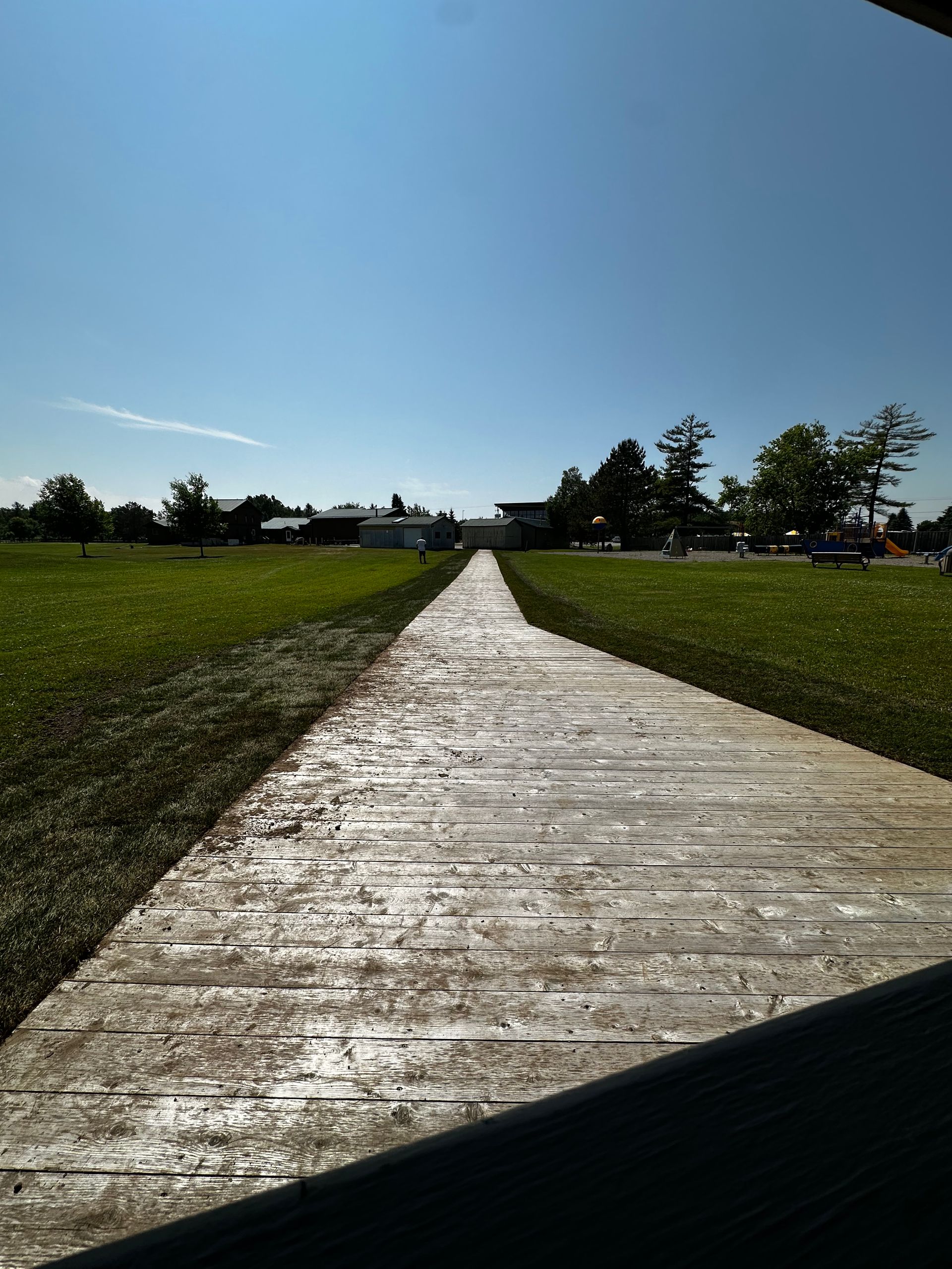 Wooden pathway through green grass under a clear blue sky, leading towards buildings.