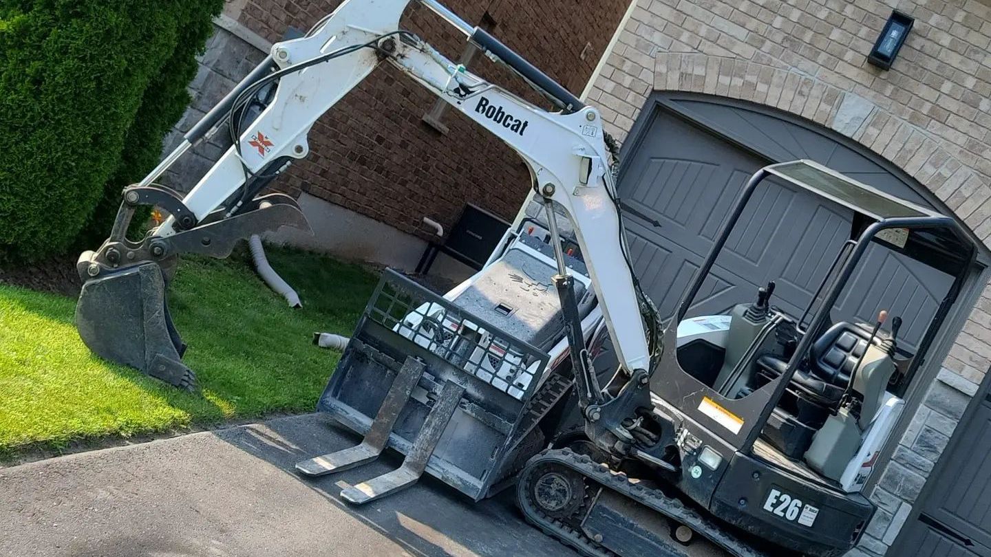 A bobcat excavator is parked in a driveway in front of a brick house.