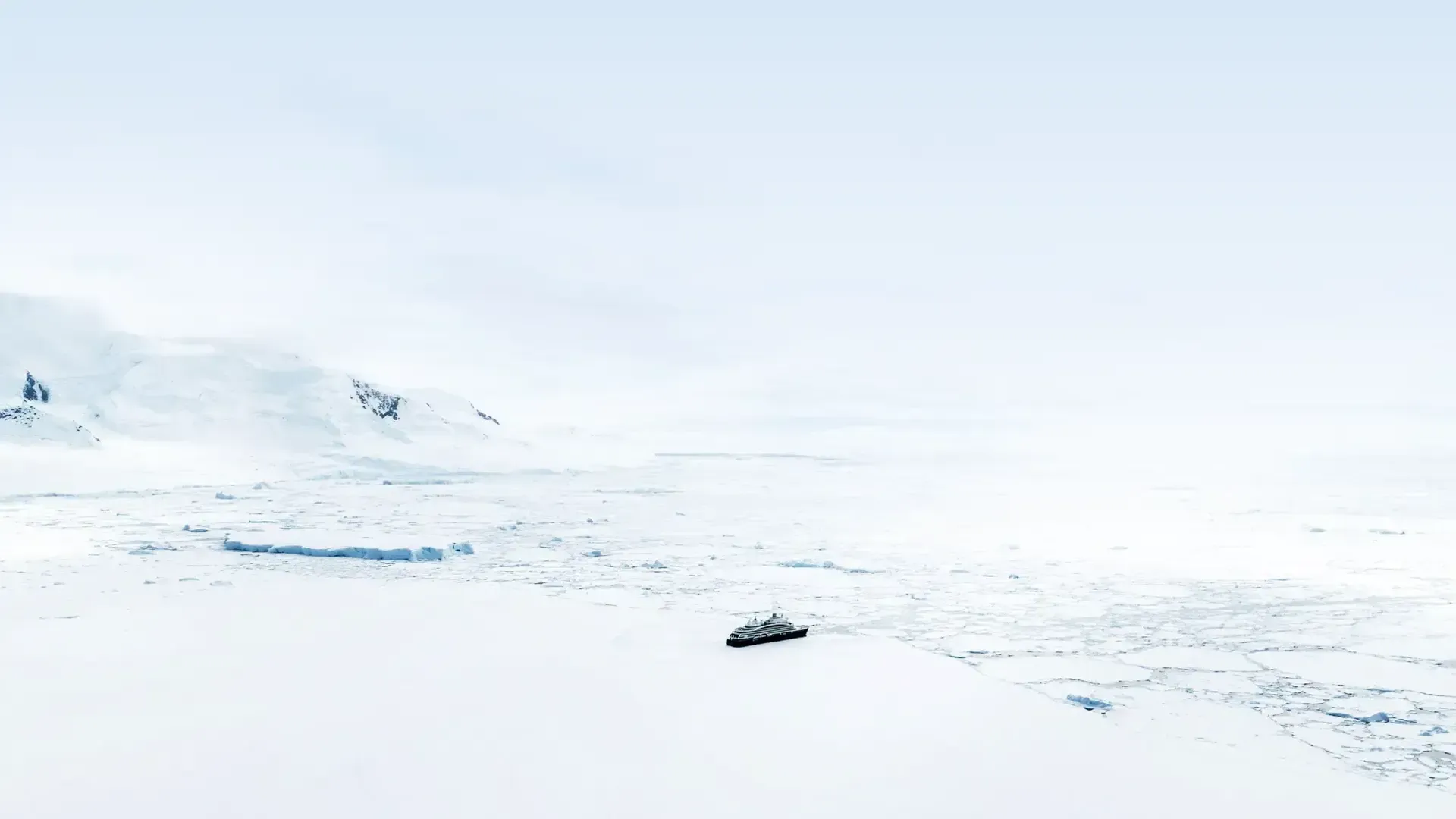 A snowy landscape with icy water and distant hills under a bright sky - Le Commandant Charcot churns its way through the ice in the center.