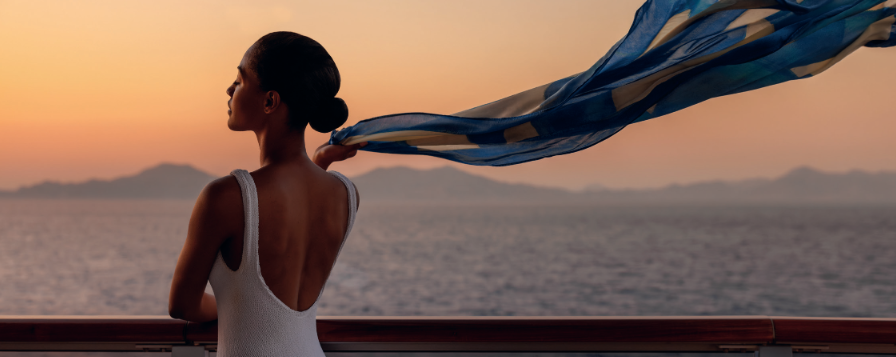 Woman in white dress by the sea, with blue scarf streaming in the wind at sunset.
