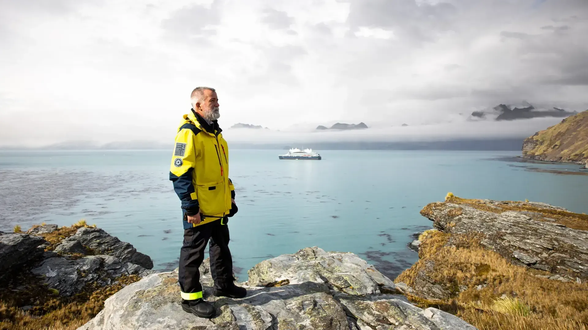 Man in a bright yellow jacket standing on rocky shore, looking over calm sea with a cruise ship in the distance