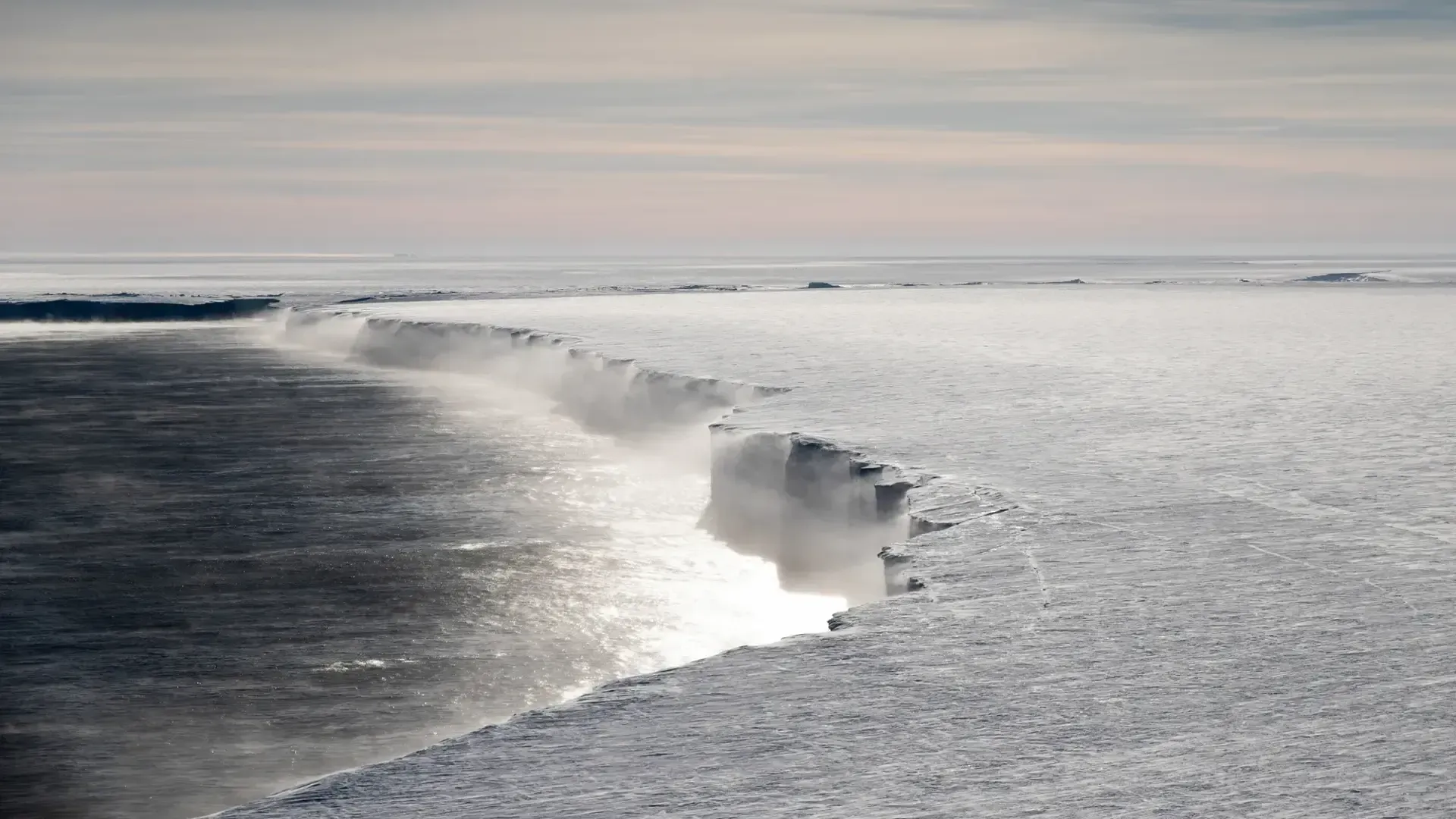 The Ross Ice Shelf stands over a deep blue sea as winds blow snow & ice off the cliffs.