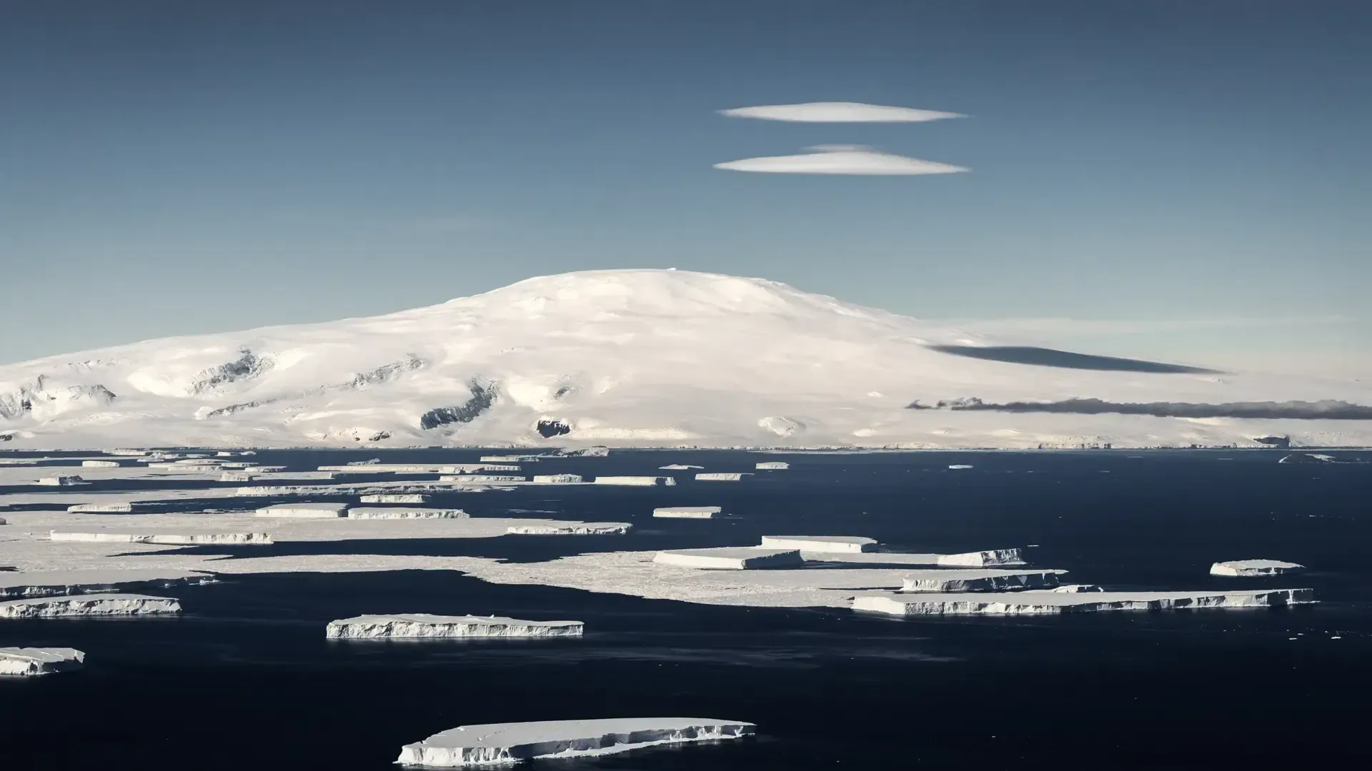 Snow-covered mountain above dark, icy water under a clear blue sky