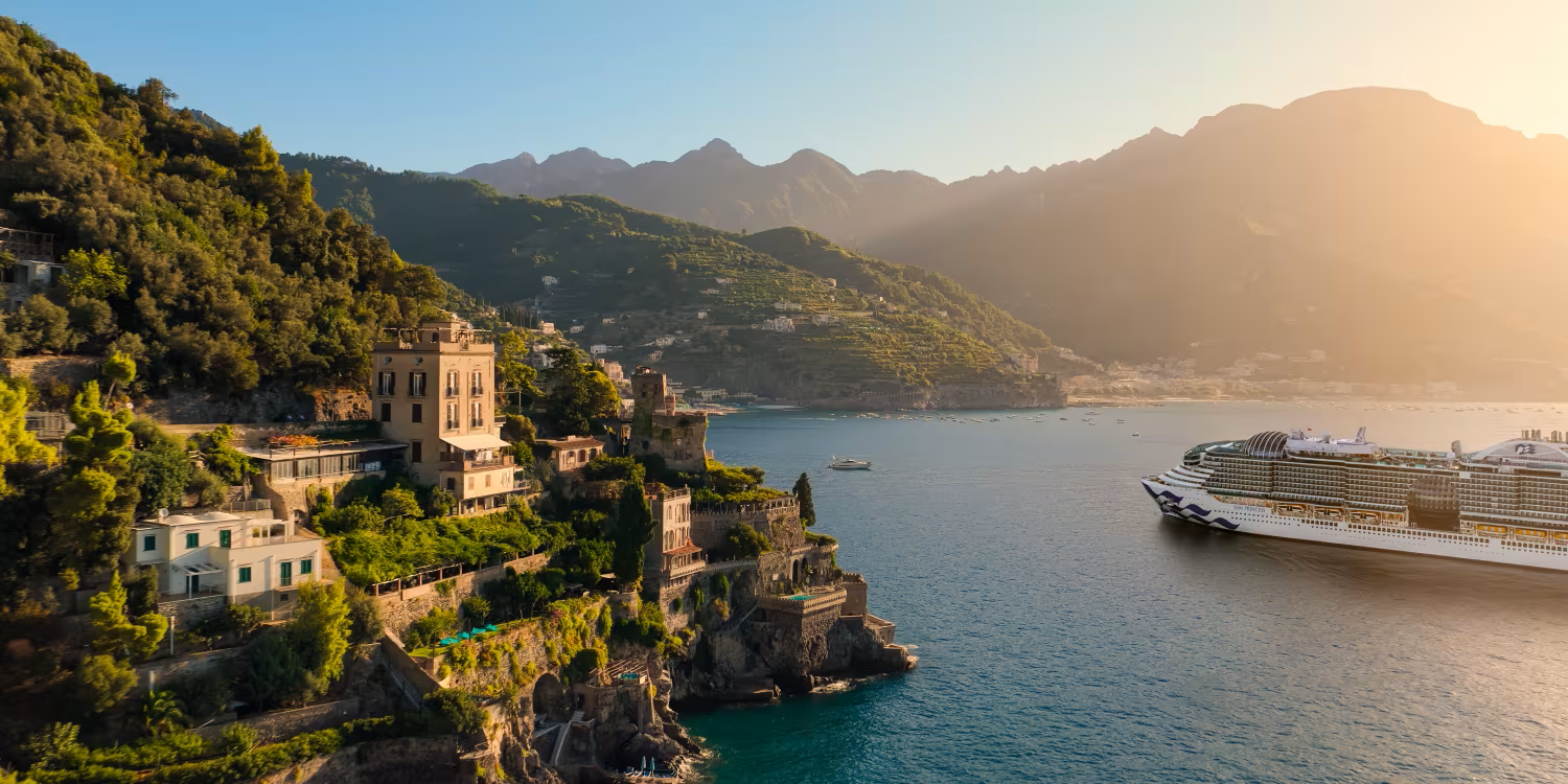 Sunlit coastal village with cliffs, boats in a bay, and mountains in the background