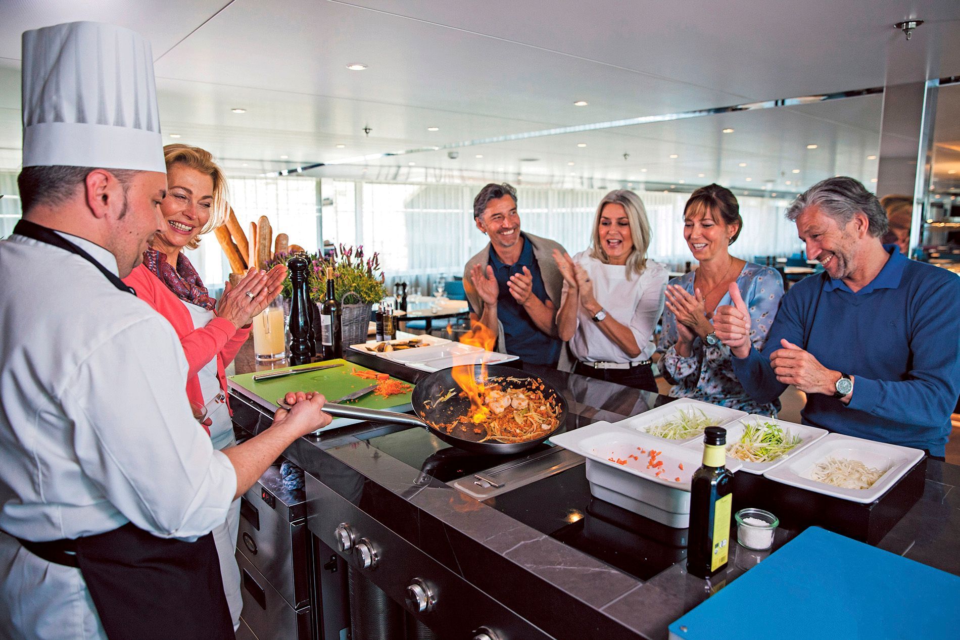 A chef demonstrates flambé cooking to a group of people at a kitchen counter, while the group watches and claps.