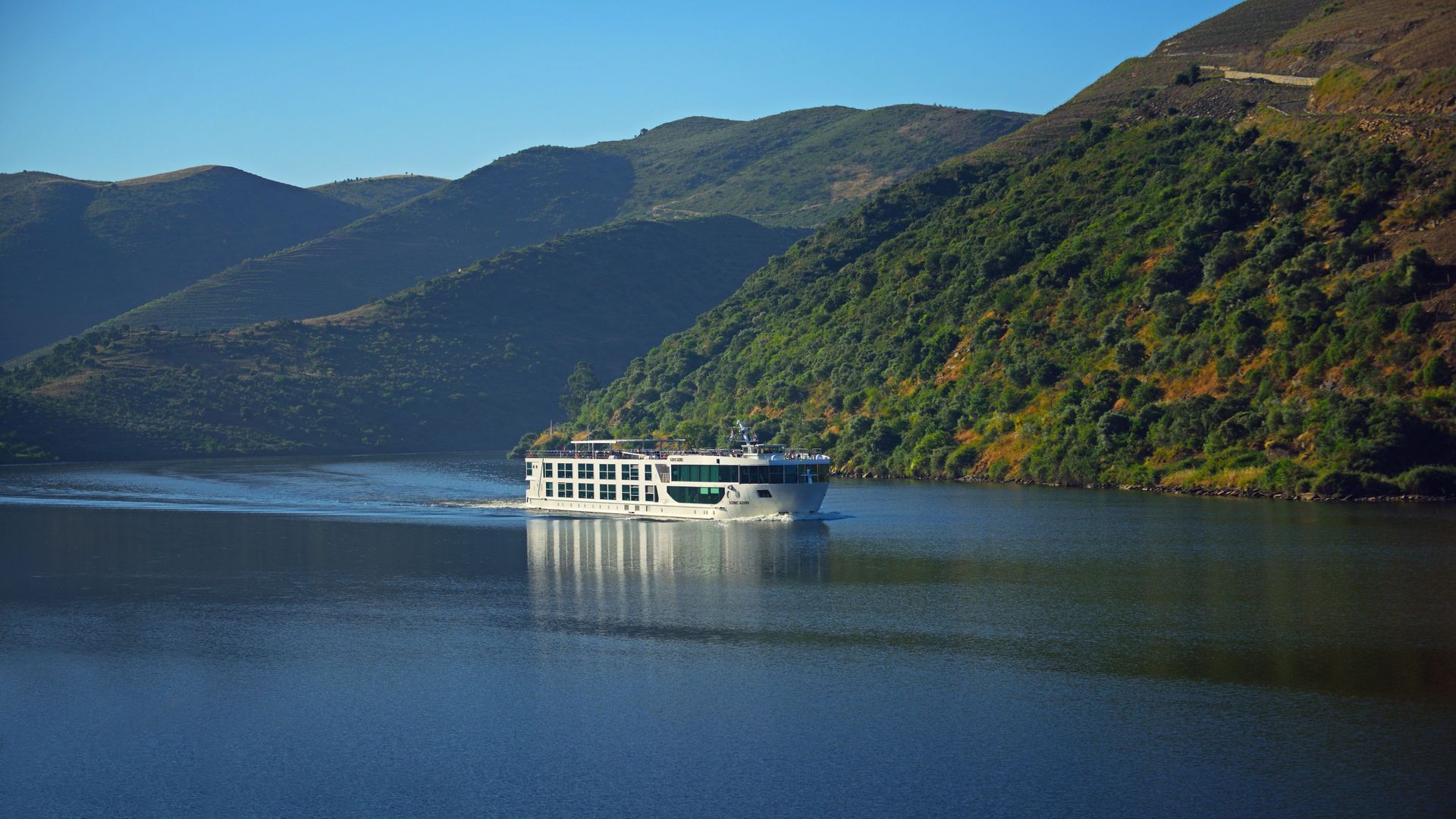 A white river cruise ship travels through a calm, wide river flanked by large, green, terraced mountains under a blue sky.