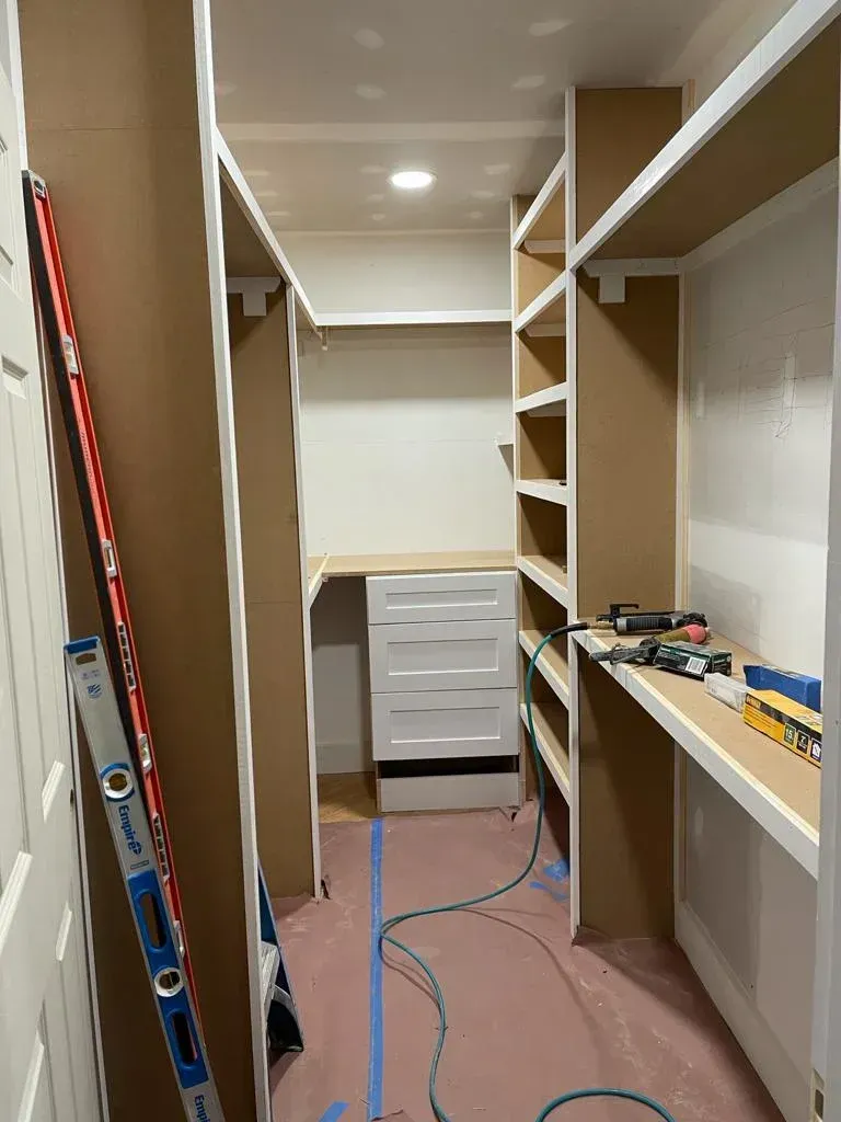 Narrow closet room with unfinished shelving, a white drawer unit, and tools on the floor.
