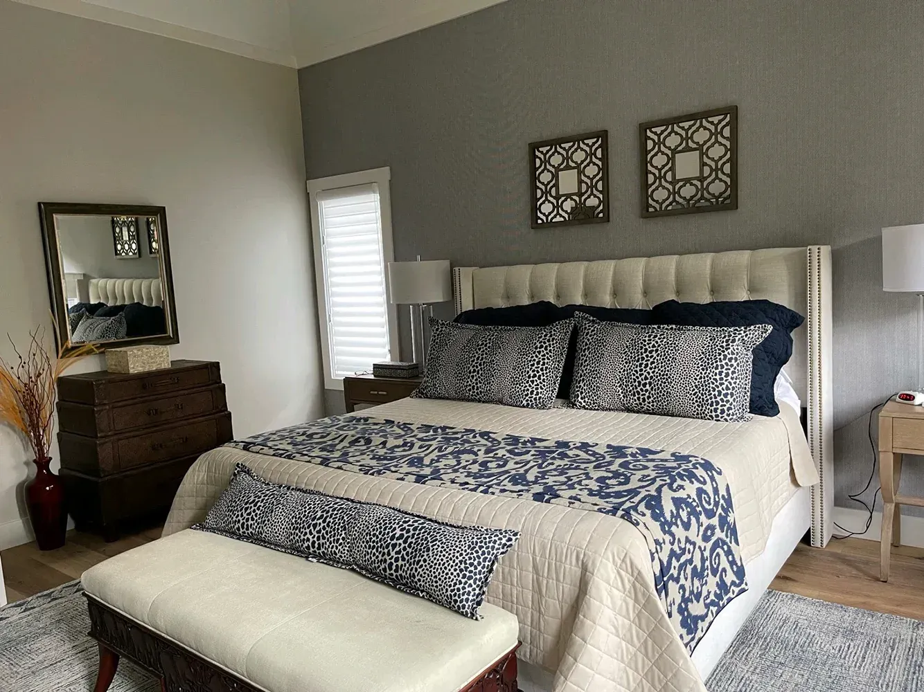 Neatly made bed with blue and white bedding in a light gray bedroom with dresser, mirror, and wall art.