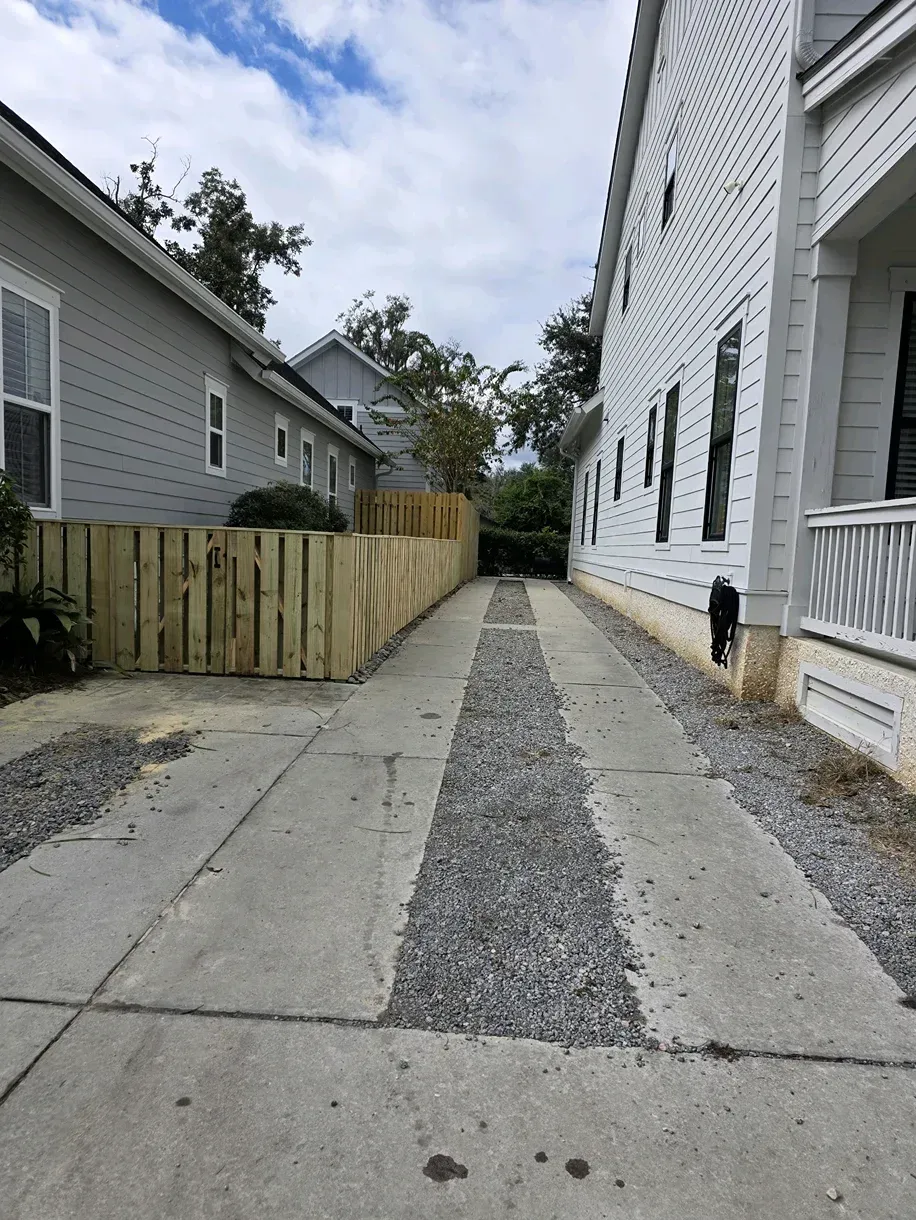 Narrow alley between two houses, with concrete and gravel path and wooden fence on the left.