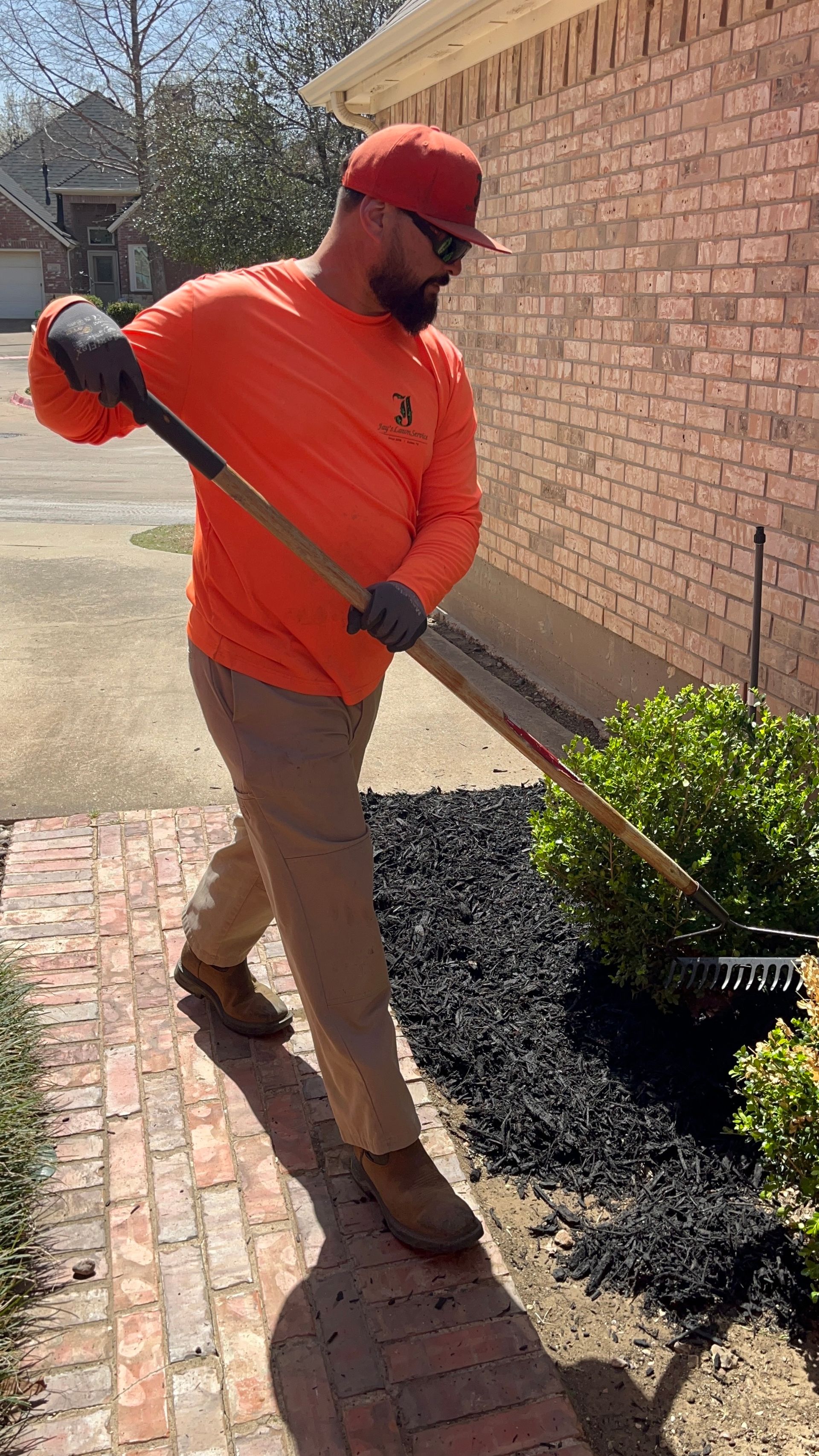 Man raking black mulch around bushes. He wears orange shirt, tan pants, hat, and gloves, near a brick building.