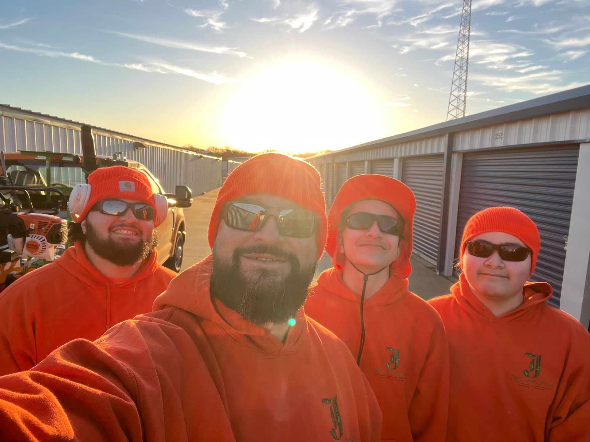 Four people in orange hoodies and hats, smiling, by storage units in the sun.