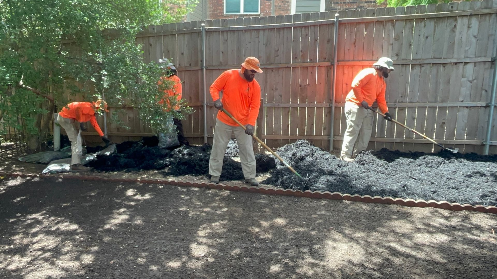 Four workers in orange shirts and hats spreading black mulch in a yard next to a fence.