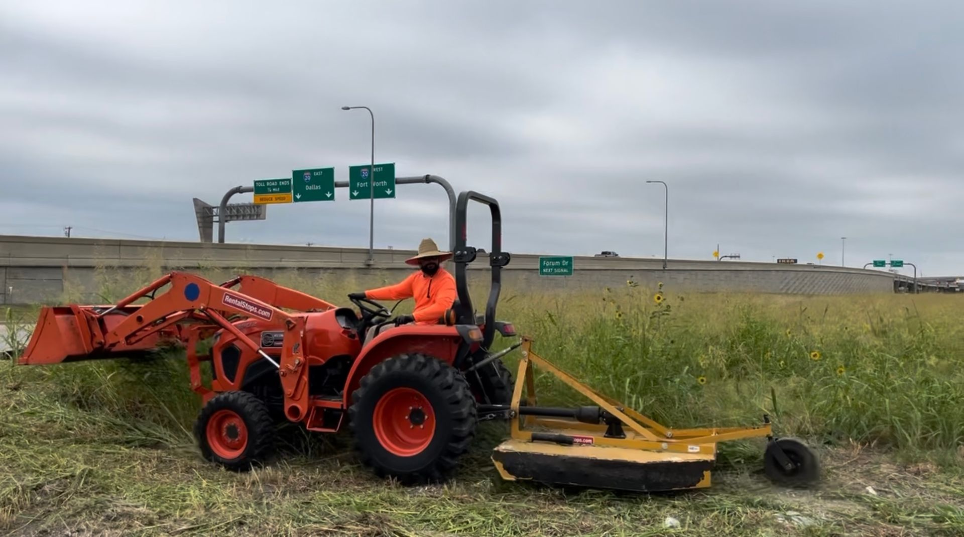 Man operating an orange tractor with a mower, cutting tall grass alongside a highway. Cloudy day.
