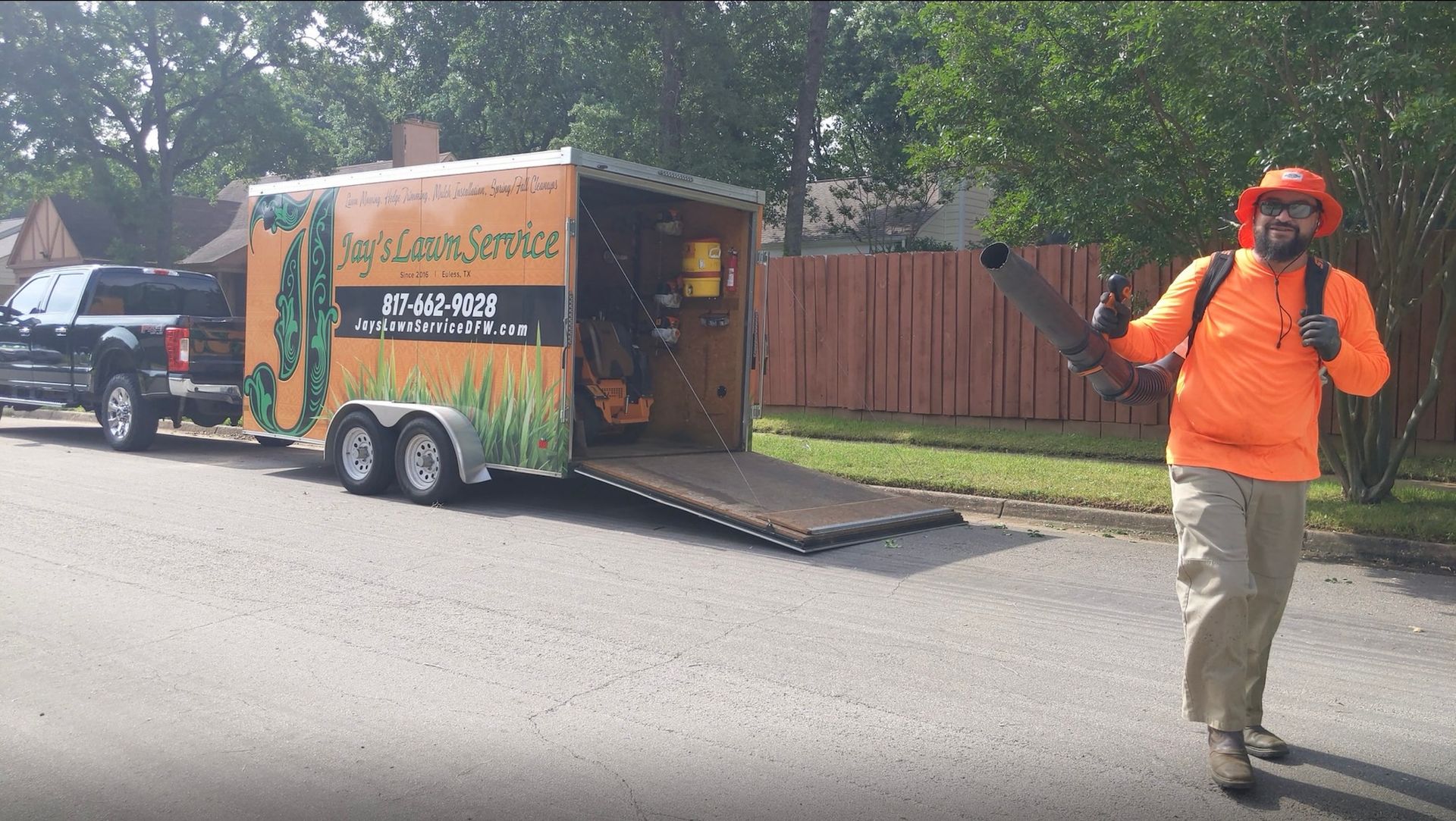 Man in orange gear with leaf blower stands next to a trailer hitched to a truck on a street.